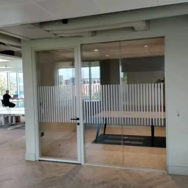 An executive office with glass partitions and doors featuring a series of circular safety dots. The room is furnished with a wooden table and chairs, and offers a view of the outside through the glass.