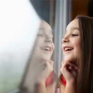 Young girl happily looking through a window, her reflection visible on the glass, emphasizing the clarity and safety provided by safety window film.