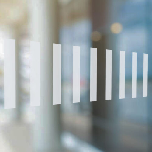 Close-up of a glass surface with a row of vertical frosted vinyl safety strips. The semi-transparent strips are evenly spaced and help improve visibility against the blurred background of an indoor commercial or office space.