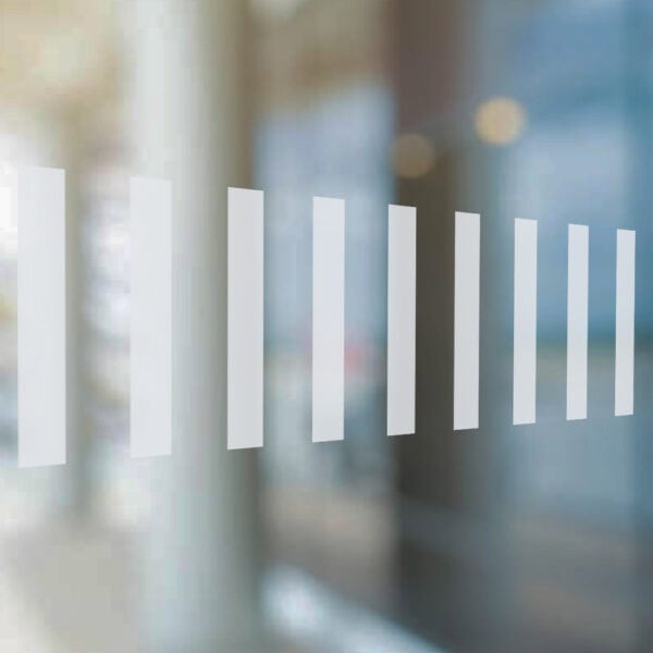 Close-up of a glass surface with a row of vertical frosted vinyl safety strips. The semi-transparent strips are evenly spaced and help improve visibility against the blurred background of an indoor commercial or office space.