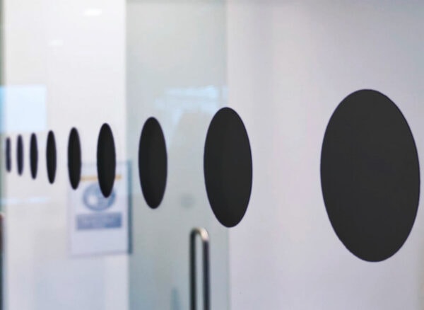 Close-up view of a glass door featuring a row of evenly spaced black vinyl safety dots. The dots appear in perspective, fading into the distance, with a modern interior environment visible through the transparent glass.