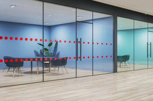 Contemporary office interior featuring glass partition walls with two horizontal rows of bright red vinyl safety dots. The transparent panels enclose modern meeting rooms with teal chairs, light wooden flooring, and soft blue-toned walls.