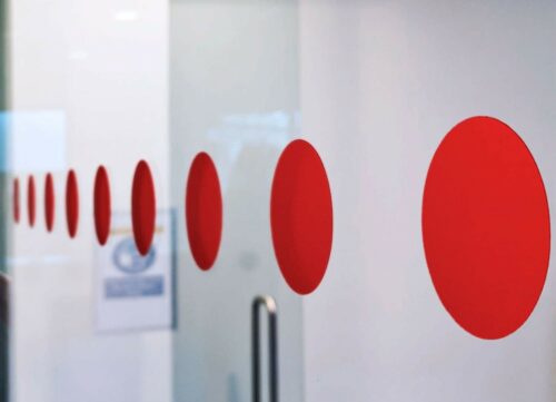 Close-up of a glass door fitted with a horizontal line of bright red vinyl safety dots. The dots appear in perspective, gradually decreasing in size as they extend into the background, against a clean, modern interior.