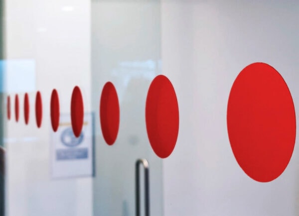 Close-up of a glass door fitted with a horizontal line of bright red vinyl safety dots. The dots appear in perspective, gradually decreasing in size as they extend into the background, against a clean, modern interior.